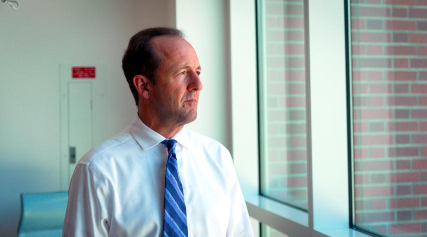 a middle aged man dressed in a white shirt and a tie looks out a hospital window with a stern expression on his face.